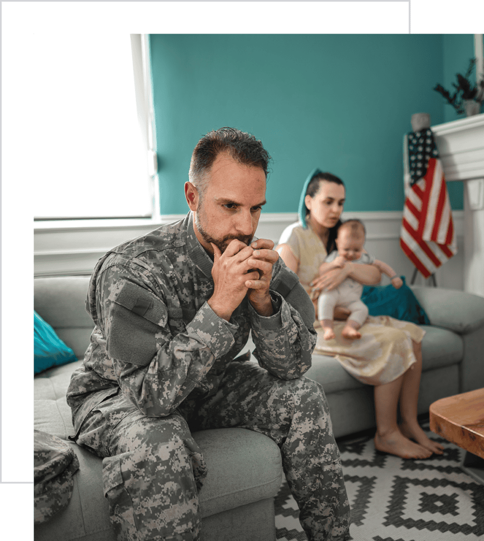 Soldier sitting pensively, family in background.